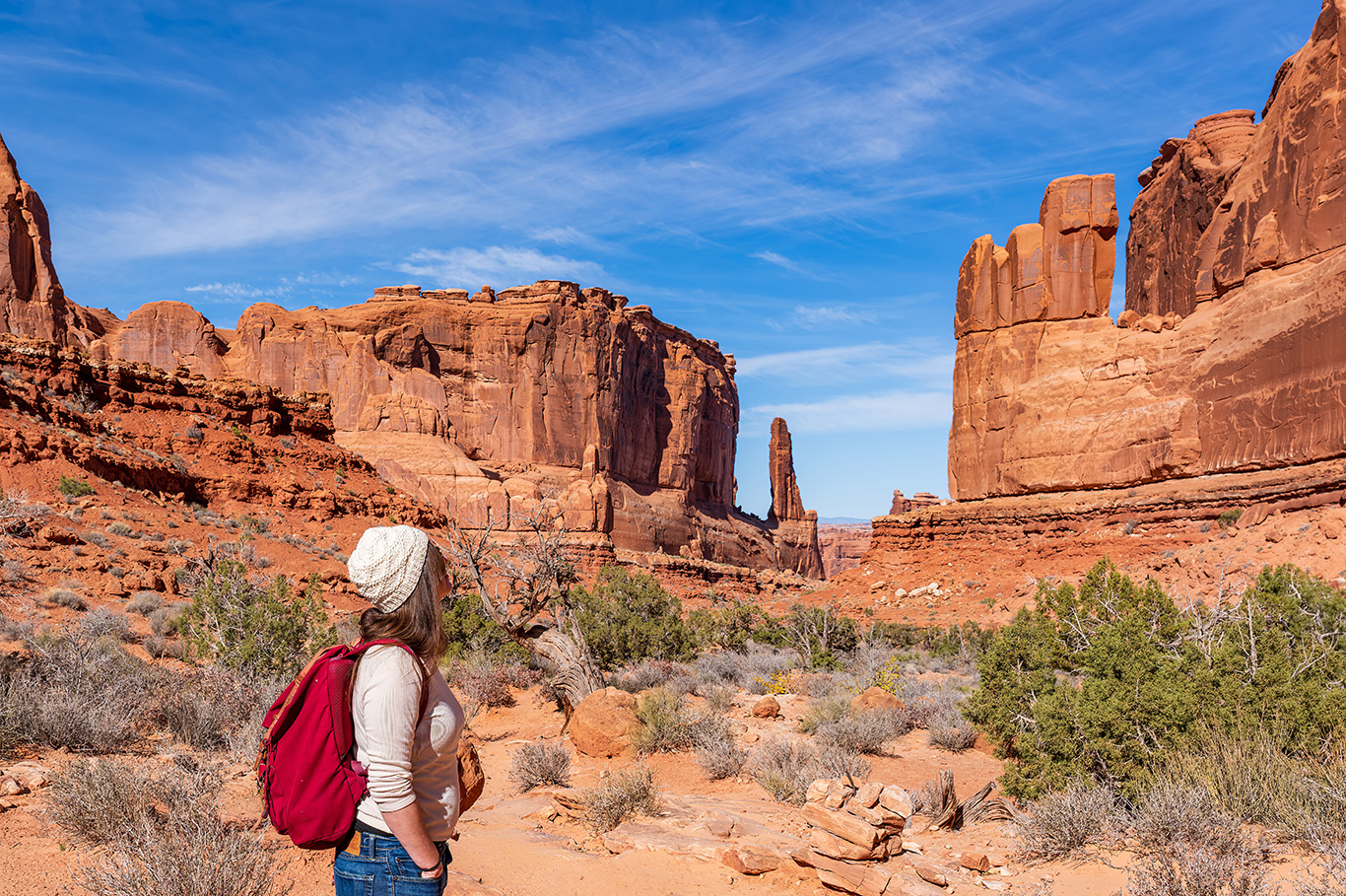 A hiker with a red backpack looking out across towering red sandstone cliffs beneath a blue sky